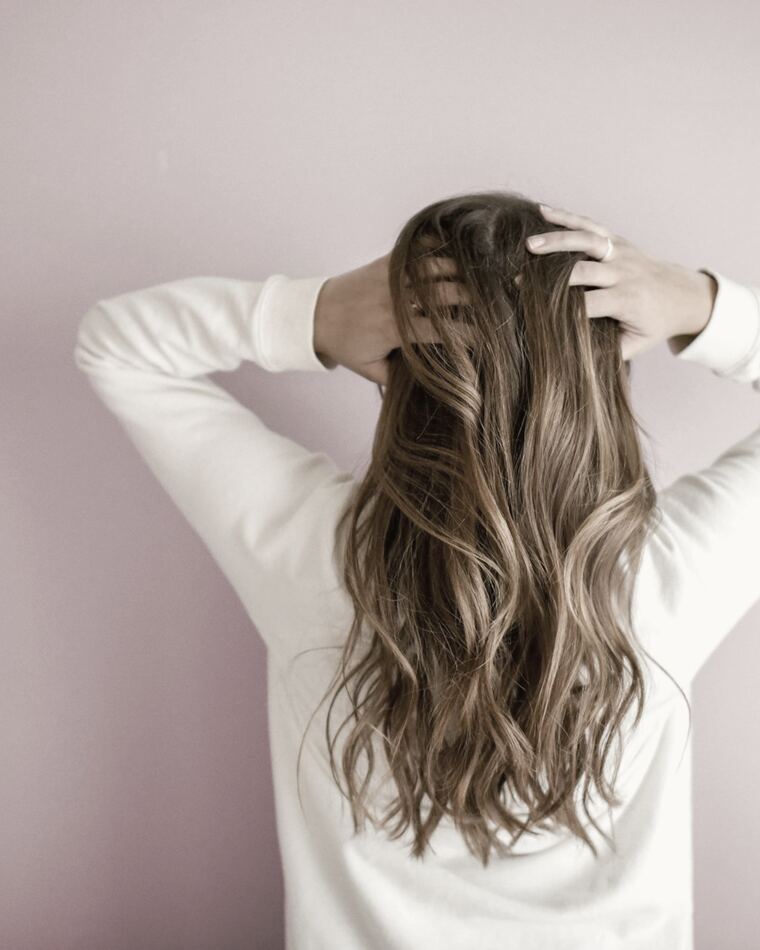 Symbolbild: Eine Frau mit langen, gewellten Haaren fährt sich mit beiden Händen durchs Haar, Rückenansicht.