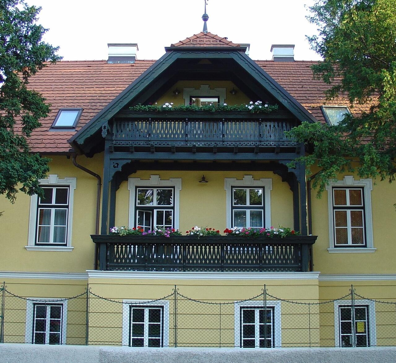 Das Haus der Schönheit am Asperner Heldenplatz 5: gelbe Villa mit rotem Ziegeldach, dunkelgrünem Holzgiebel und zwei Balkonen mit Blumenkästen.