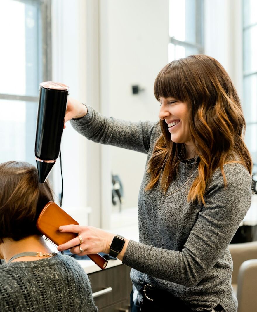 Symbolbild: Eine Friseurin föhnt und bürstet einer Kundin den Kurzhaarschnitt in einem hellen Salon.